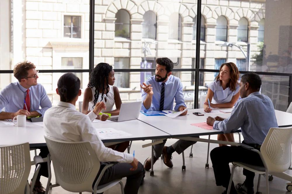 6 people around an office table talking during a business meeting.