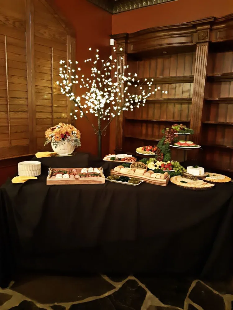 A table with a black table cloth and a barren tree with white decorative lights. Various meat, bread, cheese and fruit platters are neatly arranged on the table.